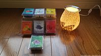 Photo showing electric wax melter lamp switched on with a lit bulb and seven packs of scented wax melts arranged behind it on the floor.