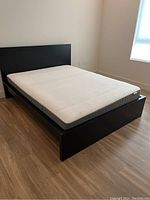 Photo shows a modern black wooden platform bed with a white mattress placed on top, positioned on a wooden floor in a well-lit room.