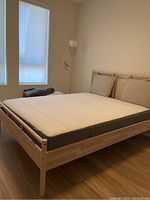 Light wood bed frame with mattress and two beige cushioned headboards in a room with wooden floor and large windows with blinds.