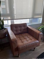 Orange-brown leather tufted armchair with well-padded seat and back cushions, set near sunny window with plants nearby.