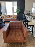 Photo of a rustic orange-brown leather chair with tufted seat in a room with other furniture.