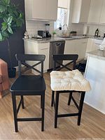Two black wooden chair stools in kitchen area, one with beige cushion on seat.