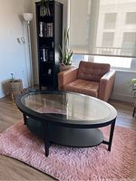 Photo showing oval dark wood coffee table with glass top and lower shelf placed on a pink rug in a living area with an armchair and bookshelf in the background.