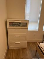 Photo showing a white wooden 4-drawer dresser with the top drawer having a frosted glass panel, placed against a wall near a window.