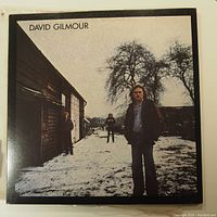 Front cover of David Gilmour self-titled vinyl LP record showing image of David Gilmour standing outside a barn or rustic building in a snowy environment.