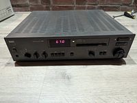 Front angle view of the NAD 7130 stereo receiver showing digital frequency display, control knobs and buttons on a light wood surface with brick wall background.