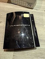 Top view of the Sony PS3 Fat console showing the PlayStation 3 embossed logo and the console's glossy black surface with scratches.