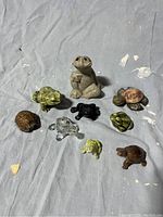 Nine assorted frog and turtle figurines displayed on a textured blue fabric background, showing various materials and colors.