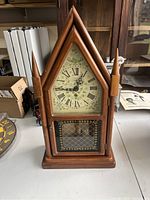 Front view of wooden antique clock with pointed arch design on a table with Roman numeral face and decorative glass panel.