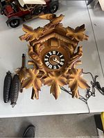 Front view of the wooden cuckoo clock showing carved birds and leaves, the clock face with Roman numerals, and two pinecone-shaped weights with chain.