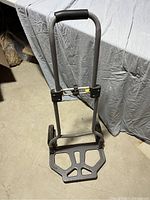 Folding hand cart standing upright with handle extended, showing metal frame, rubber grip, and plastic base platform.