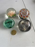 Photo showing four glass paperweights on white table with a coin for size reference. Paperweights include a white flower, a millefiori star, a green seed pod, and dandelion seed heads inside clear glass.