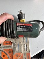 Close-up of Bosch PWS 620-S angle grinder label showing model, power, and speed specs with coiled power cord and plug.