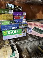 Photo showing stacked puzzle boxes including Golden Guld 1500-piece puzzle, Puzzle Roll Away mat in pink, and several 1000-piece puzzles with varying artwork including forest and landscapes.