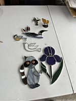 Photo of four stained glass decorative items on a white table: two birds (one in blue/yellow, one hummingbird), a raccoon with amber eyes, and a purple iris flower with green leaves. The raccoon shows signs of needing repair.