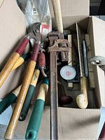 Top view of a box containing garden shears, pipe wrench, and wooden handled hand tools showing wear and rust