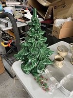 Green ceramic Christmas tree with electrical cord, displayed on white table among various other household items. Clear plastic bag with red and green bulbs placed in front.