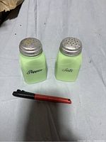 Two green milk glass salt and pepper shakers placed side by side on a white cloth surface with a pen for scale.