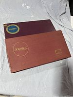 Two vintage Scrabble game boxes, one maroon with blue and gold logo, the other reddish-brown with gold lettering and publisher logo, placed side by side.