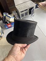 Hand-held view of black top hat with brown ribbon trim on a table.