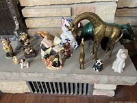 Photo showing mixed animal figurines on stone fireplace ledge including large brass giraffe, white ceramic rabbit, multicolored ceramic birds, several small bears and dogs.
