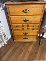 Front view of tall wooden dresser showing five drawers with ornamental brass handles and worn surface.