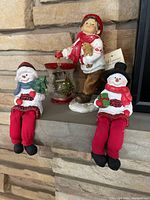 Photo showing two snowman figurines sitting with red pants and a child figurine standing, one snowman holding a tree, the child holding a lantern and teddy bear