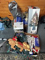Wide view showing boxed artificial tree, multiple boxes of light strings, and bag with decorations including wooden Santas and reindeer
