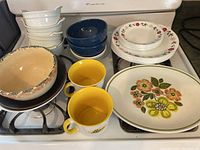 Overview of various dishes on stove including bowls, floral plates, large mixing bowl, and Leggs mugs
