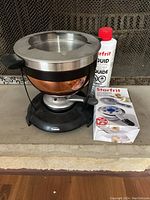 Stainless steel copper exterior fondue pot with black burner base, bottle of Starfrit fuel liquid and burner box on fireplace hearth.