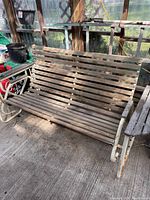 Photo of a wooden garden swing with horizontal slats and white metal frame inside a greenhouse or enclosed porch, showing wear and dust.