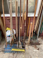 Full view of assortment showing rakes, hoes, picks, axes, shovels, and brooms with wooden handles and metal parts leaning against a wall.