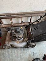 Full side view of gas lawn mower showing metal deck, wheels, and attached fabric bagger in need of cleaning.