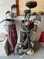 Two golf bags filled with men's left hand golf clubs. One bag is red Spaulding with irons and woods, the other is black and gray on a pull cart.