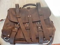 Front view of brown leather computer or travel bag showing three buckle straps securing the front flap and handle on top.