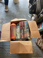 Photo showing open cardboard box with colorful plastic straws in container, two packaged BBQ lighters, folded tarp, and other smaller items inside the box on garage floor.