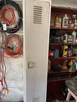 Photo of the closed tall, narrow metal locker with vents in the door, painted light grey, against a wall with hanging cables nearby.