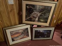 Set of three framed prints, one held up showing garden with water wheel, rocky seashore and duck pond prints stacked nearby.
