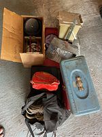 Overview of rock tumbler box, metal toolboxes, ropes and putty knife on concrete floor with signs of use and dirt.