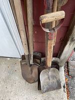 Garden shovels with wooden handles and metal blades showing heavy use and some rust, stored leaning against wall.