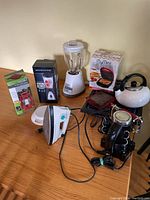 Full view of all items on wooden table including blender, iron, sandwich maker, hand mixer, griddle, kettle, and lantern unpackaged.