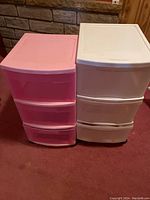 Photo showing front view of pink and white plastic storage units with three drawers each on carpeted floor.