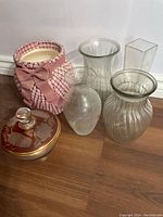 Five different vases plus a decorative glass container with lid arranged on wooden floor.