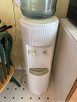 Full view of white water cooler with large empty water jug on top, positioned between chair and counter on tiled floor.
