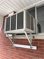 Large white window air conditioner installed in a brick window frame, mounted on metal support brackets outdoors.