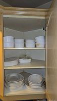 Corning Ware bakeware stored inside wood cabinet, three shelves visible. Top shelf has 6 smaller oval and round dishes, middle shelf has 3 dishes, bottom shelf has larger oval and round dishes stacked.