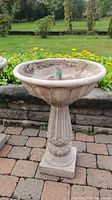 Complete weathered two-piece concrete birdbath outdoors on brick patio with garden background, viewed from front