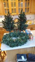 Two small prelit artificial Christmas trees and artificial Christmas wreath displayed on a kitchen table with some decorations visible on trees.