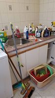 Photo showing countertop with multiple cleaning liquid bottles, flat mop, brooms, and basket with cleaning brushes