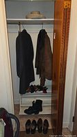 Closet view with two coats hanging and shoes/boots on racks below, plus a straw hat on upper shelf.